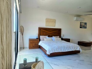 A bedroom with a large bed, wooden headboard, bedside tables, a wall-mounted air conditioner, and a window with curtains.