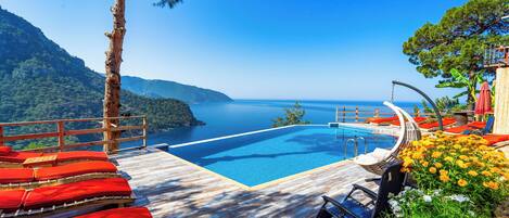 A poolside area with wooden deck, red lounge chairs, and a view of the sea.