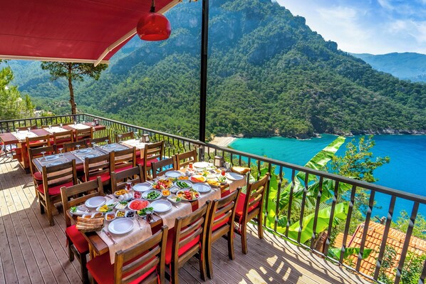 A terrace with tables set for dining, overlooking a mountainous coastline.