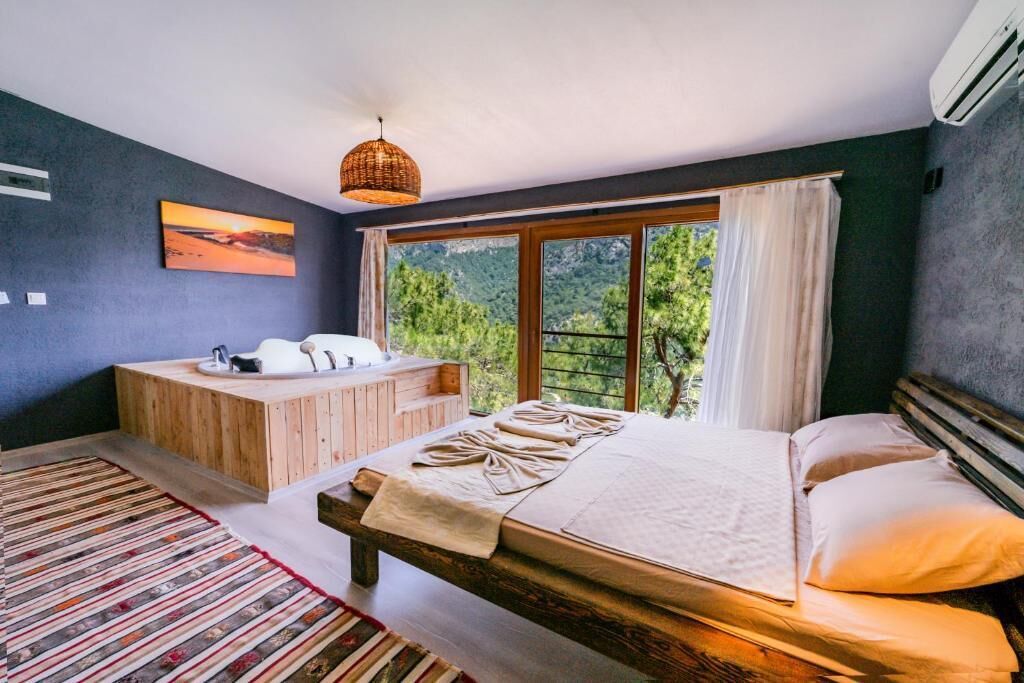 A bedroom with a large bed, a wooden bench, a bathtub, and a view of greenery through a window.