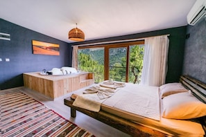 A bedroom with a large bed, a wooden bench, a bathtub, and a view of greenery through a window.