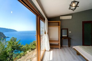 A room with a large window overlooking a coastal landscape, a bed, a wooden shelf, and a hanging light fixture.