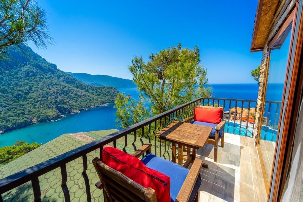 Un balcon avec une table et des chaises en bois, donnant sur un paysage montagneux et un plan d’eau.