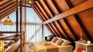 A high-ceilinged room with a sloped wooden ceiling, a bed with a white bedspread, a black chair, and a hanging light fixture.
