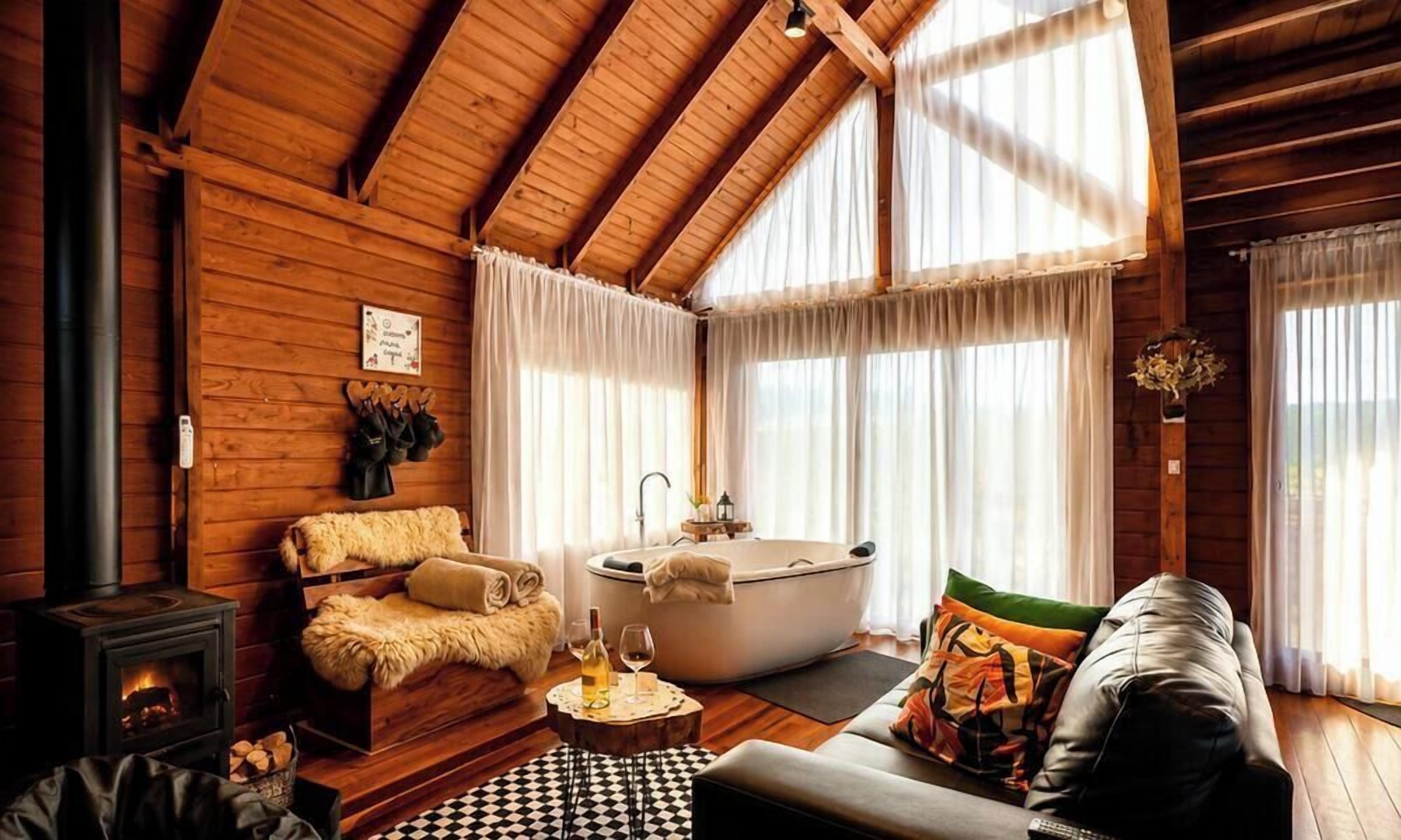 A log cabin interior with a fireplace, a sofa, a bathtub, and a large window with curtains.