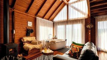 A log cabin interior with a fireplace, a sofa, a bathtub, and a large window with curtains.