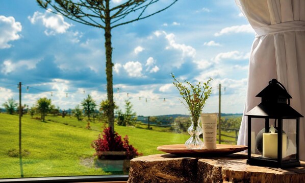 A view from a window with a wooden table holding a candle, a glass, and a vase with greenery.