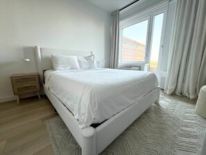 A bedroom with a bed, a nightstand, a window with curtains, and a view of buildings outside. - 10 Min Soho NYC Comfort Suite (Jersey City)