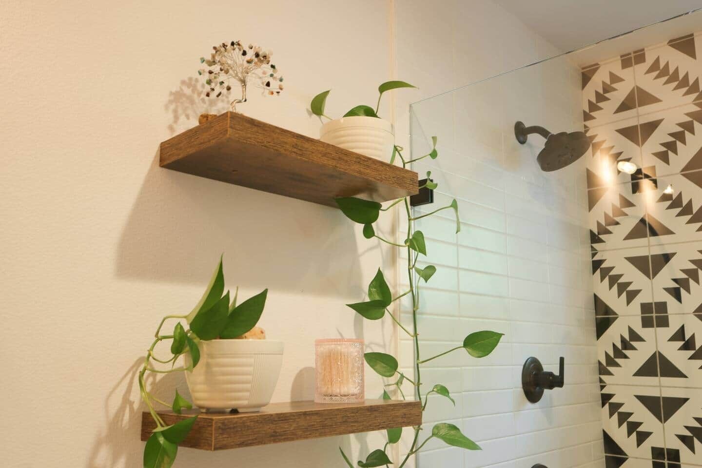 A bathroom with wooden shelves holding plants and a decorative tree, white tiled walls, and a geometric-patterned shower door.