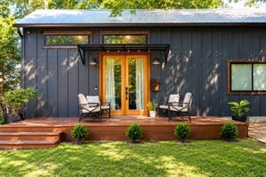 A modern house with a dark exterior, a wooden deck, two chairs, and a glass door. - Mid-Century Tiny House (Austin)
