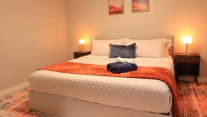 A neatly made bed with a white duvet and orange blanket, two pillows, and a rolled towel on the bed. - Retro Oasis (Austin)