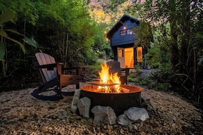 A cozy outdoor seating area with a fire pit, surrounded by rocks and greenery, and a small blue house with a lit window.