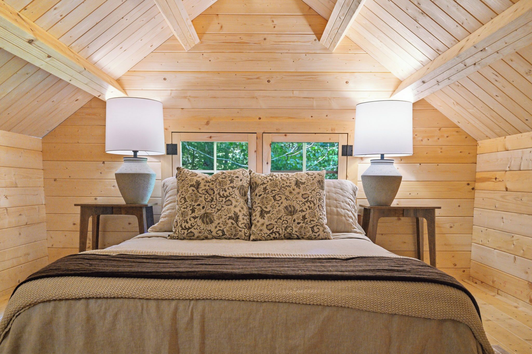 Un dormitorio acogedor con techo de madera, una cama con almohadas estampadas y dos lámparas de noche.