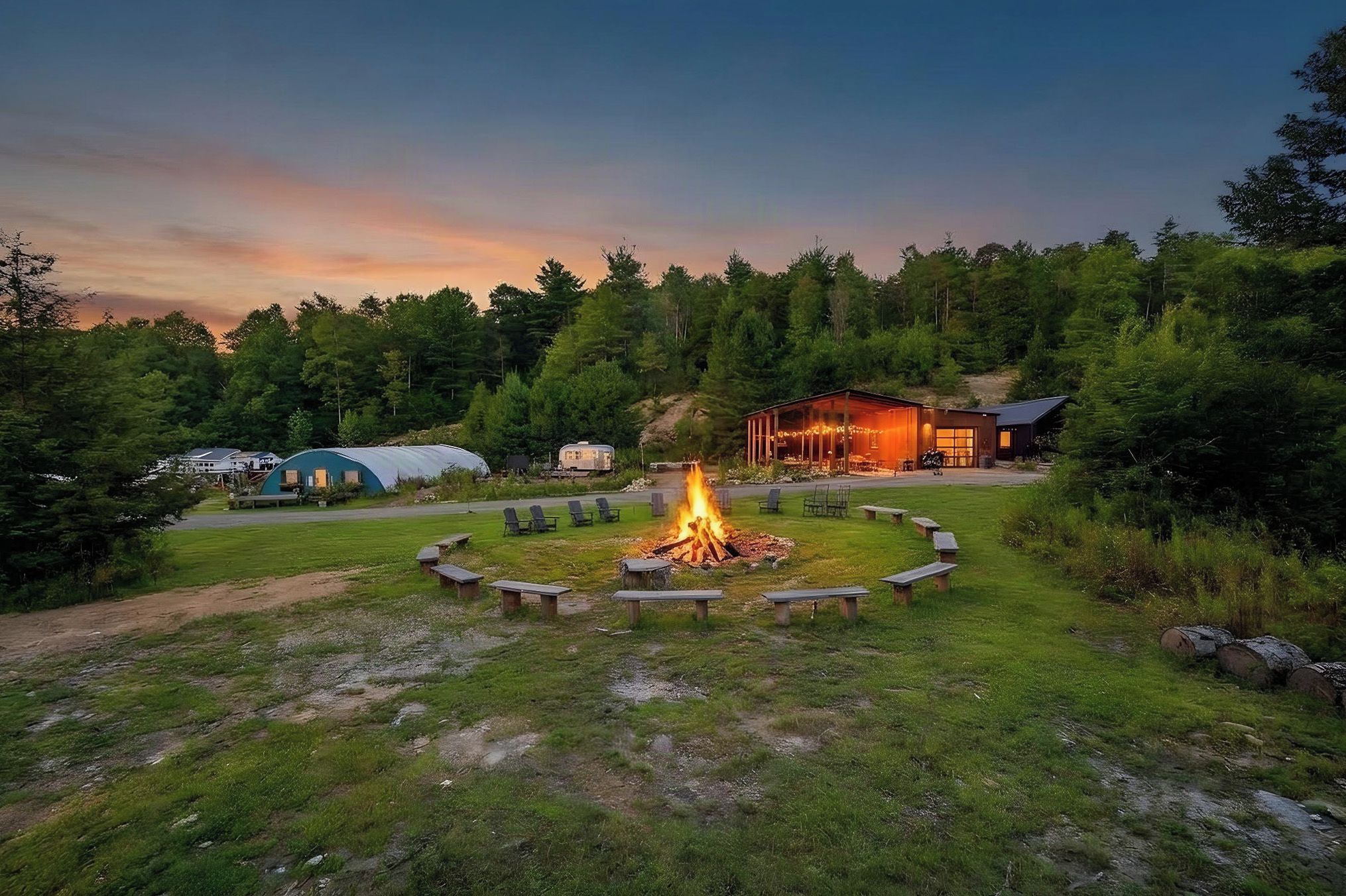 A campsite with a central fire pit surrounded by benches, a covered structure, and a building with a porch.