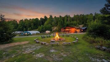 A campsite with a central fire pit surrounded by benches, a covered structure, and a building with a porch.