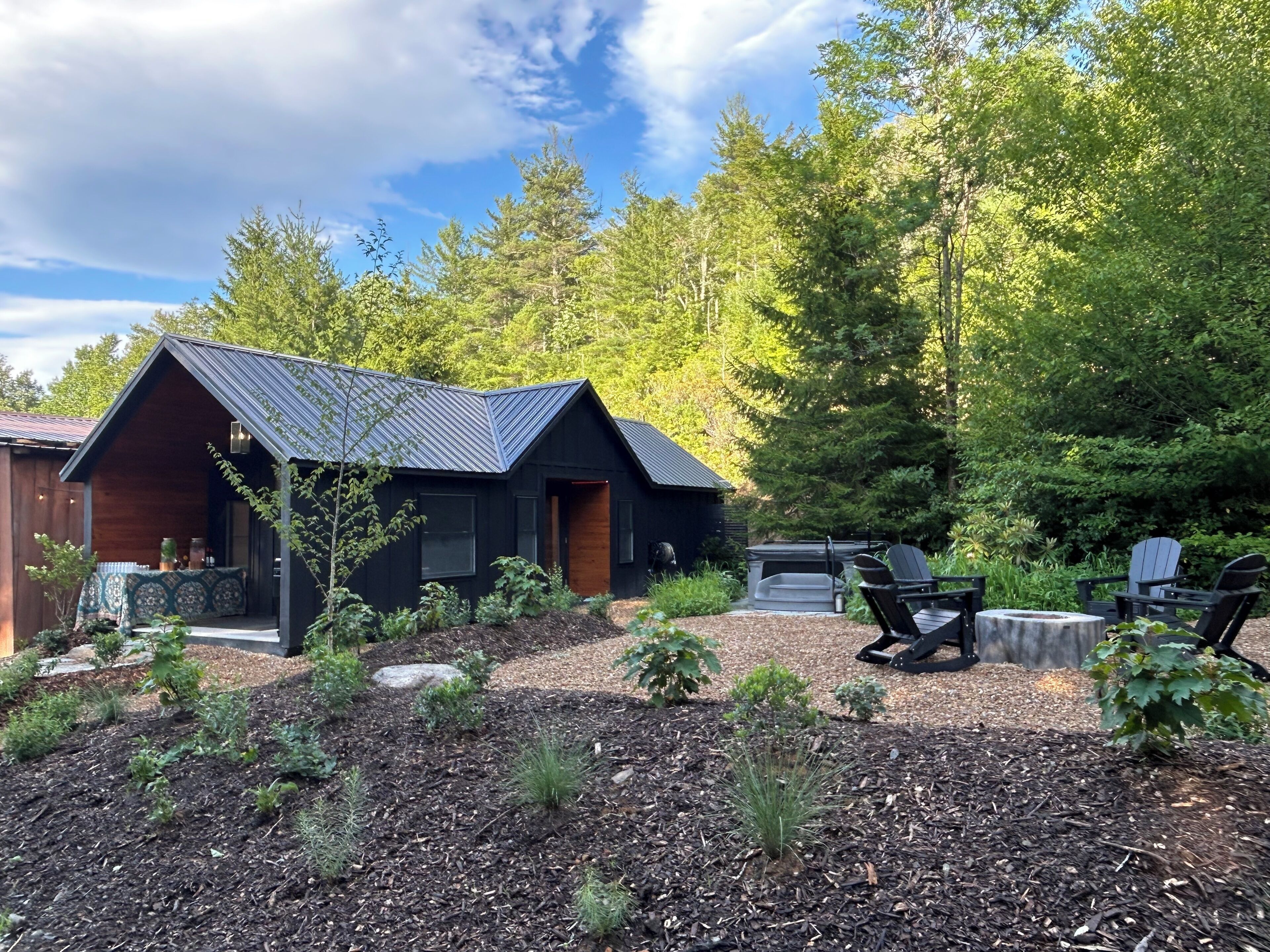 A cabin with a sloped roof, surrounded by a garden with various plants and a barbecue area.