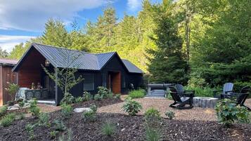 A cabin with a sloped roof, surrounded by a garden with various plants and a barbecue area.