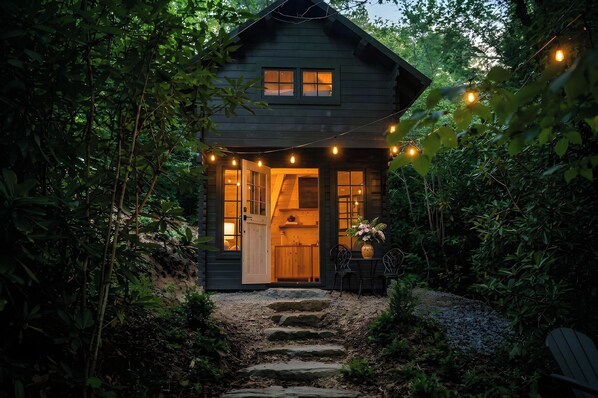A small, dark wooden cabin with a stone staircase leading to the entrance, surrounded by dense foliage.