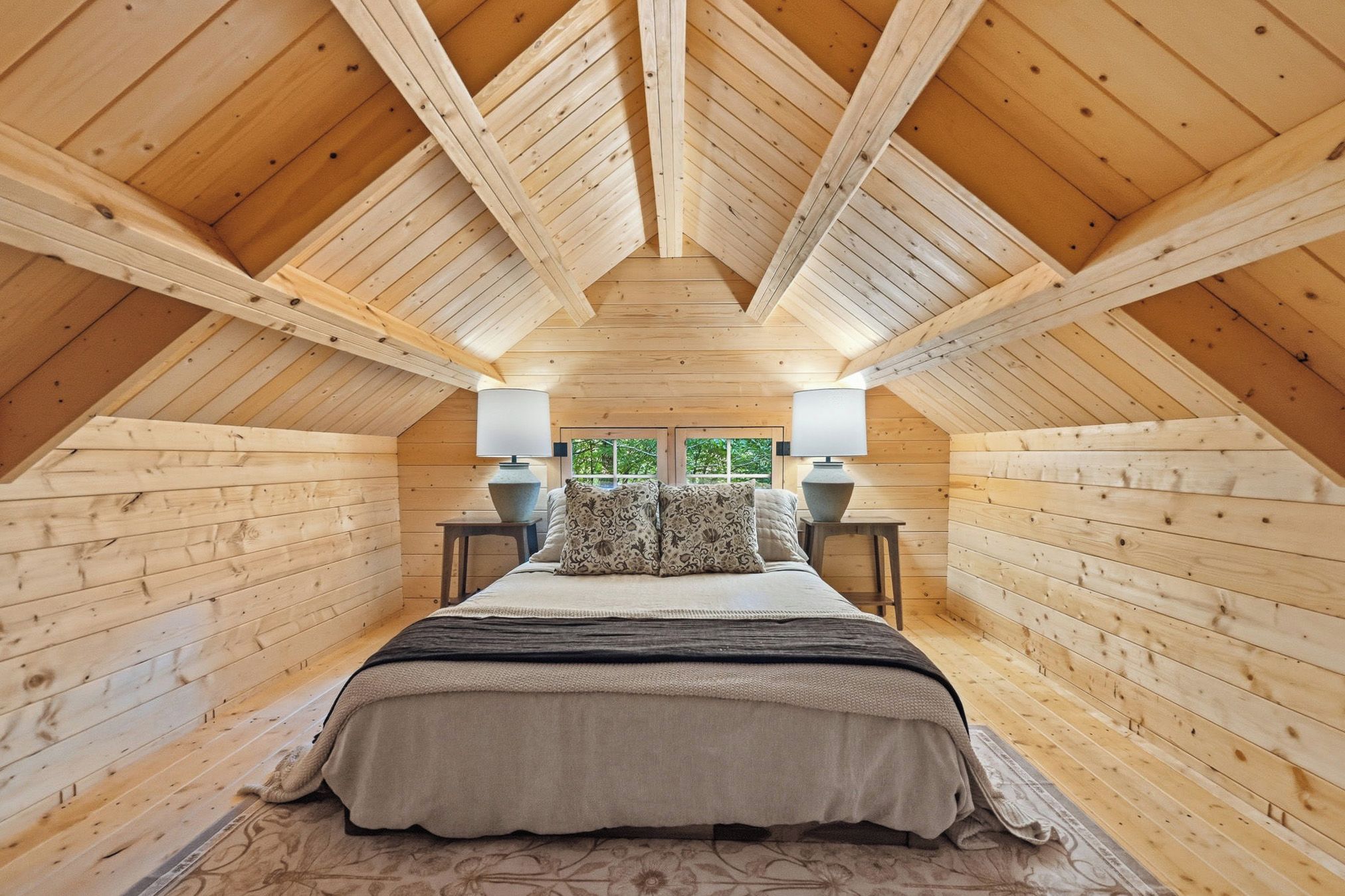 A bedroom with a sloped ceiling, wooden walls, a bed with patterned pillows, bedside lamps, and a view through a window.