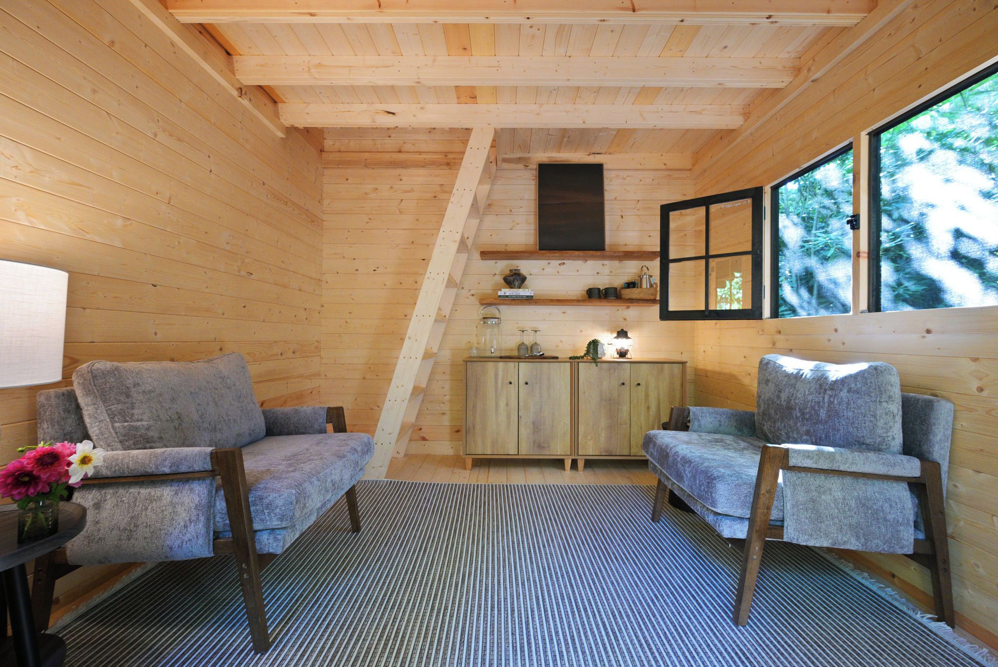 A cozy cabin interior with two upholstered chairs, a wooden cabinet, and a window with a view of trees.