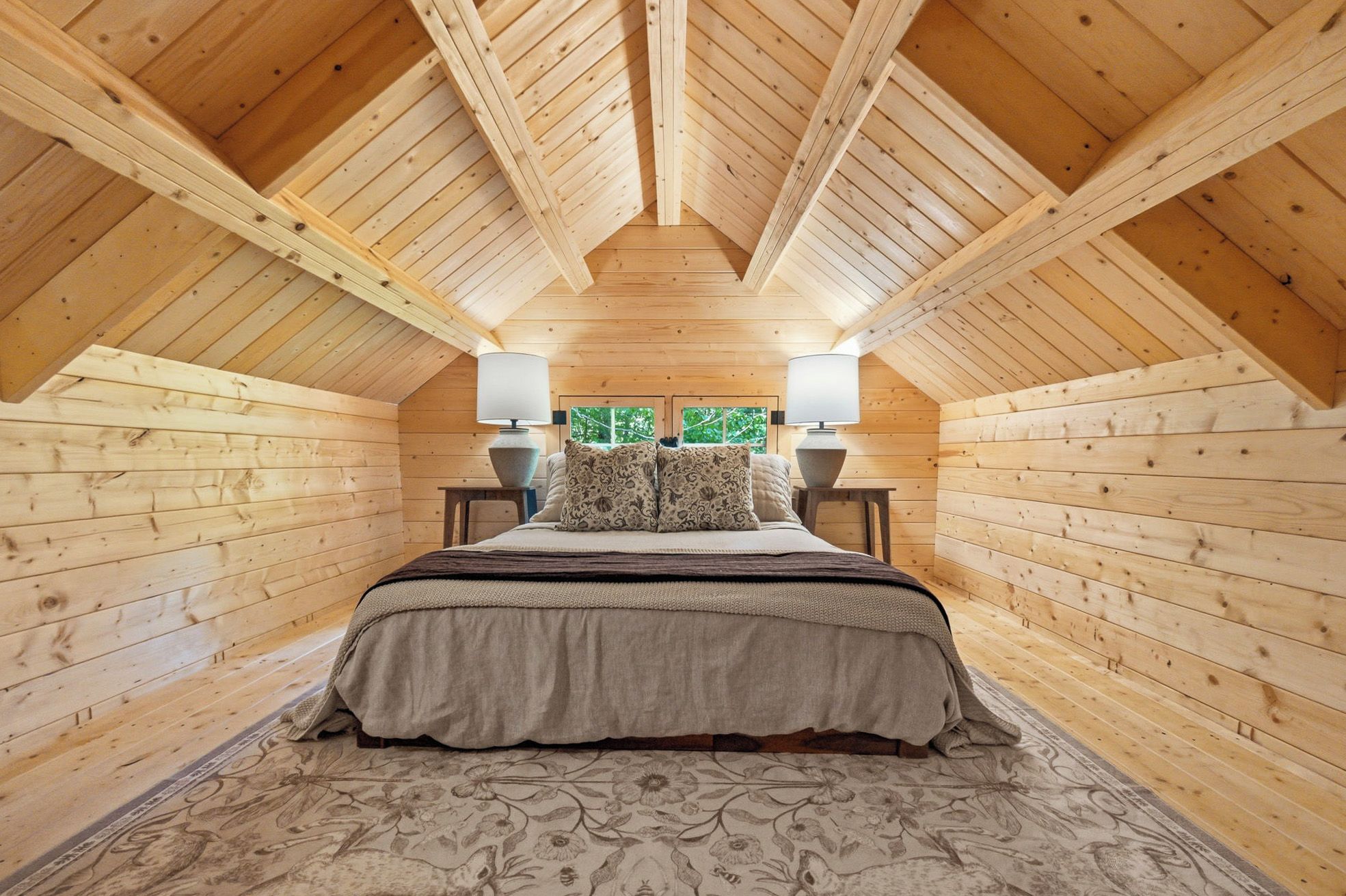 Ein Schlafzimmer mit Holzboden, einem Bett mit gemustertem Überwurf, zwei Nachttischlampen und Blick auf Grünfläche außerhalb.