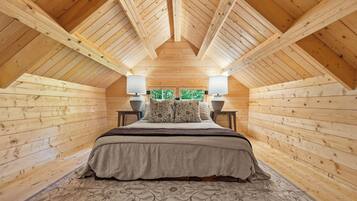 Ein Schlafzimmer mit Holzboden, einem Bett mit gemustertem Überwurf, zwei Nachttischlampen und Blick auf Grünfläche außerhalb.