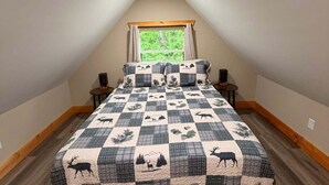 A bedroom with a checkered bedspread featuring animal prints, a window with curtains, and a sloped ceiling. - Whispering Timber 1-bedroom cabin in charming Old Fort with AC (Old Fort)