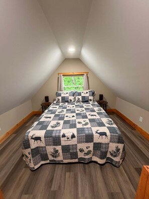 A bedroom with a checkered bedspread featuring animal prints, a window with curtains, and a sloped ceiling. - Whispering Timber 1-bedroom cabin in charming Old Fort with AC (Old Fort)