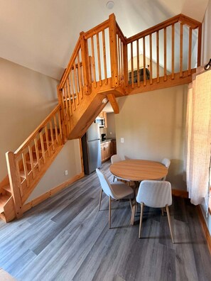 A loft-style room with a wooden staircase, a dining area with a round table and chairs, and a kitchen in the background. - Whispering Timber 1-bedroom cabin in charming Old Fort with AC (Old Fort)