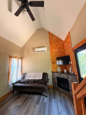 A cozy living room with a sofa, a fireplace, a ceiling fan, and a TV mounted on the wall. - Whispering Timber 1-bedroom cabin in charming Old Fort with AC (Old Fort)