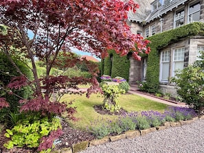 Un jardín bien cuidado con diversas plantas y un sendero de piedra que lleva a un edificio de fachada de piedra y una ventana.