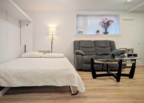 A bedroom with a bed, a gray armchair, a small table, a window with blinds, and a wall-mounted lamp.