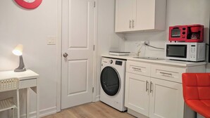 A compact kitchen with a washing machine, microwave, and a red clock on the wall. - Modern 1-Bedroom Apartment with AC in Flushing (FLUSHING)