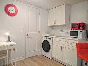 A compact kitchen with a washing machine, microwave, and a red clock on the wall. - Modern 1-Bedroom Apartment with AC in Flushing (FLUSHING)