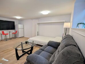 A modern room with a bed, a sofa, a small table, a TV, and a refrigerator. - Modern 1-Bedroom Apartment with AC in Flushing (FLUSHING)