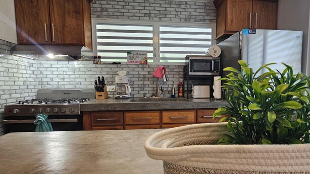 A kitchen with a gas stove, microwave, and stainless steel refrigerator. There is a sign on the window, a potted plant, and wooden cabinets.