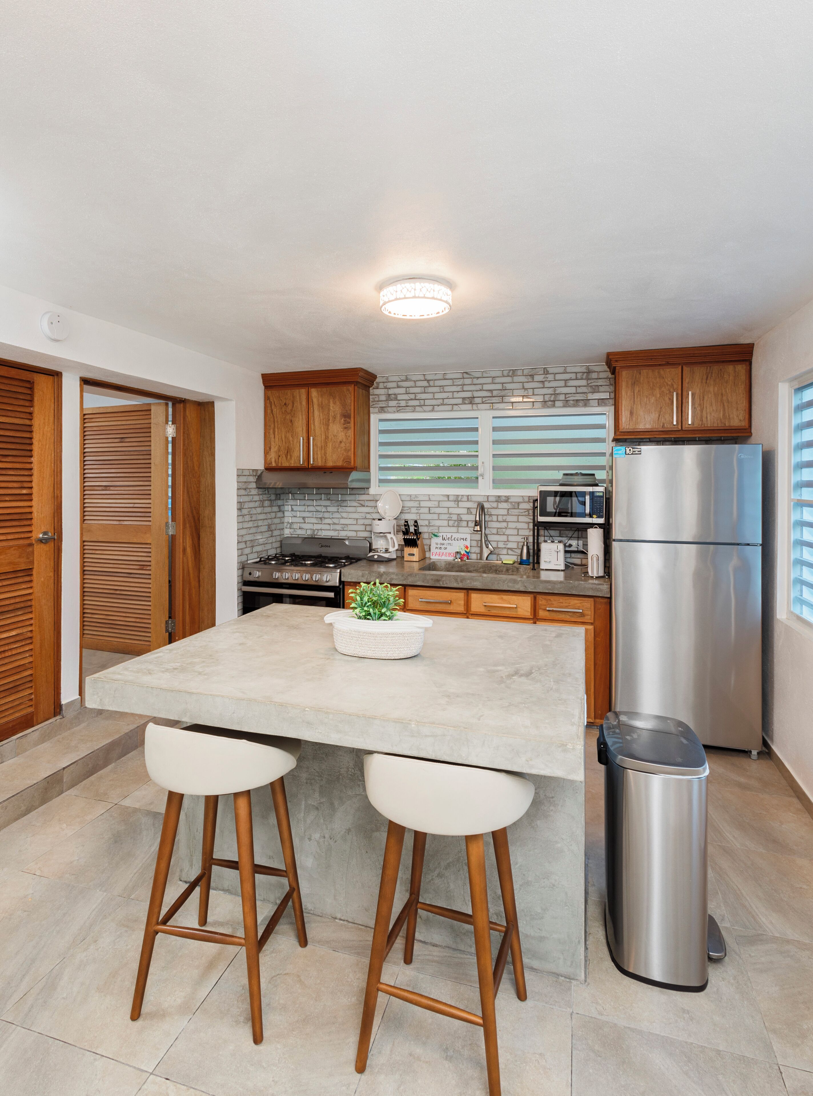 A modern kitchen with a central island, wooden cabinets, a stainless steel refrigerator, and a trash can.