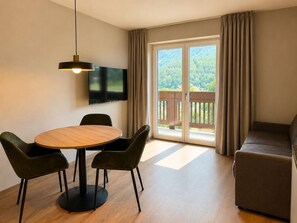 Ein modernes Wohnzimmer mit einem runden Holztisch, zwei grünen Polstersesseln, einem dunklen Sofa, einem flachbildfernseher an der Wand und einer Glastür, die auf einen Balkon mit Blick auf ein bewaldetes Gebiet führt.