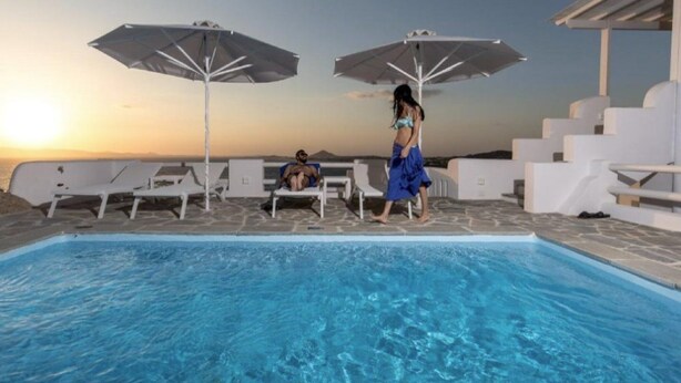 Une piscine avec des chaises longues et des parasols, une personne qui se promène et un coucher de soleil en arrière-plan.