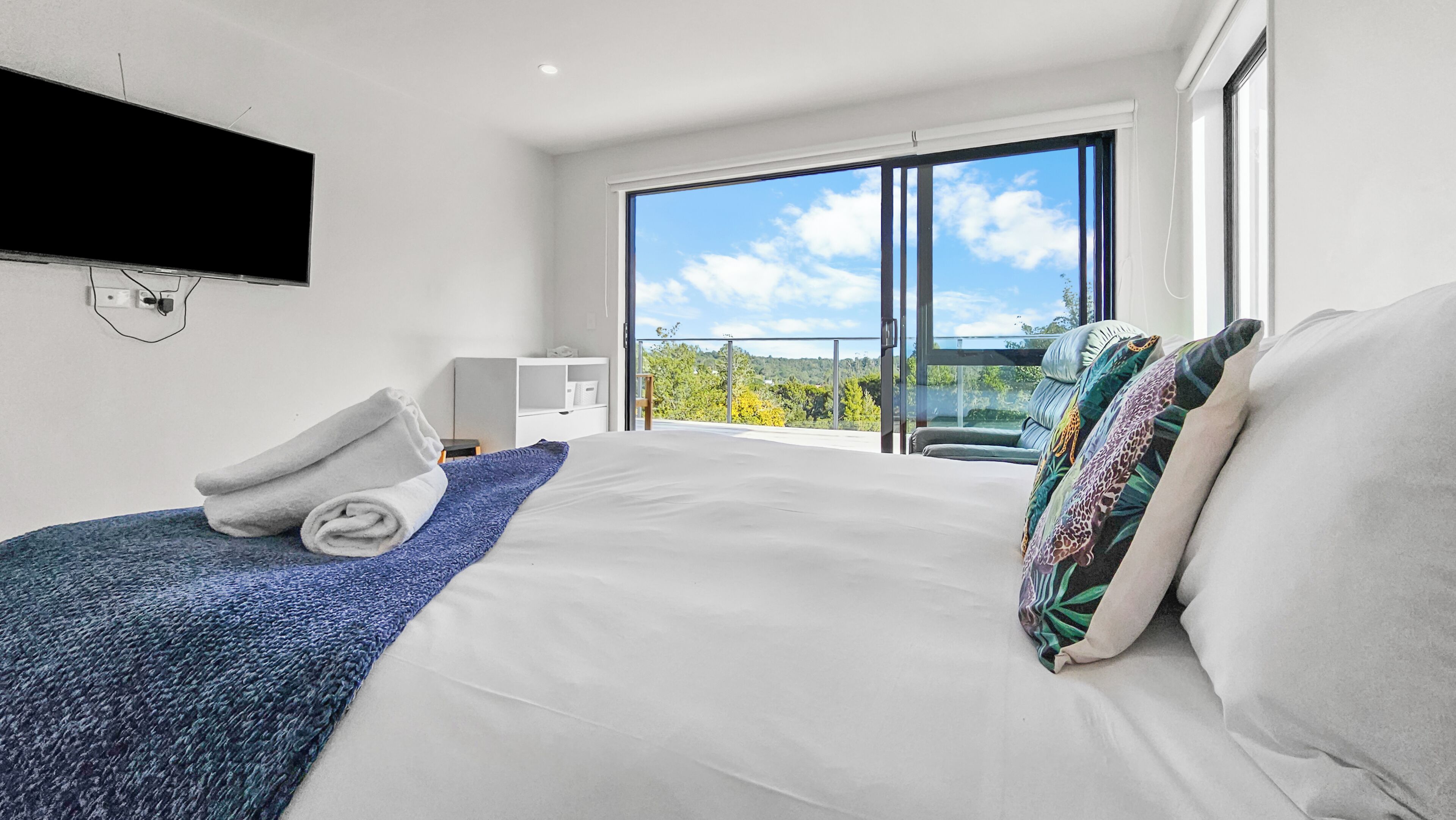 Une chambre d’hôtel moderne dotée d’un grand lit, d’un téléviseur à écran plat fixé au mur, d’un balcon offrant une vue sur le ciel et les arbres, ainsi que d’un bureau avec une chaise.