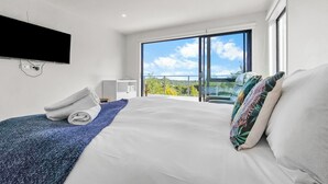 A modern hotel room with a large bed, a flat-screen TV mounted on the wall, a balcony with a view of the sky and trees, and a desk with a chair. - Birkenhead Bliss | Serene 4Bed Home | Quiet, Views (Birkenhead)