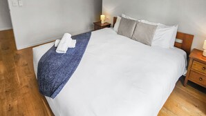A bedroom with a large bed, wooden flooring, a nightstand with a lamp, and a wall-mounted light. - Birkenhead Bliss | Serene 4Bed Home | Quiet, Views (Birkenhead)