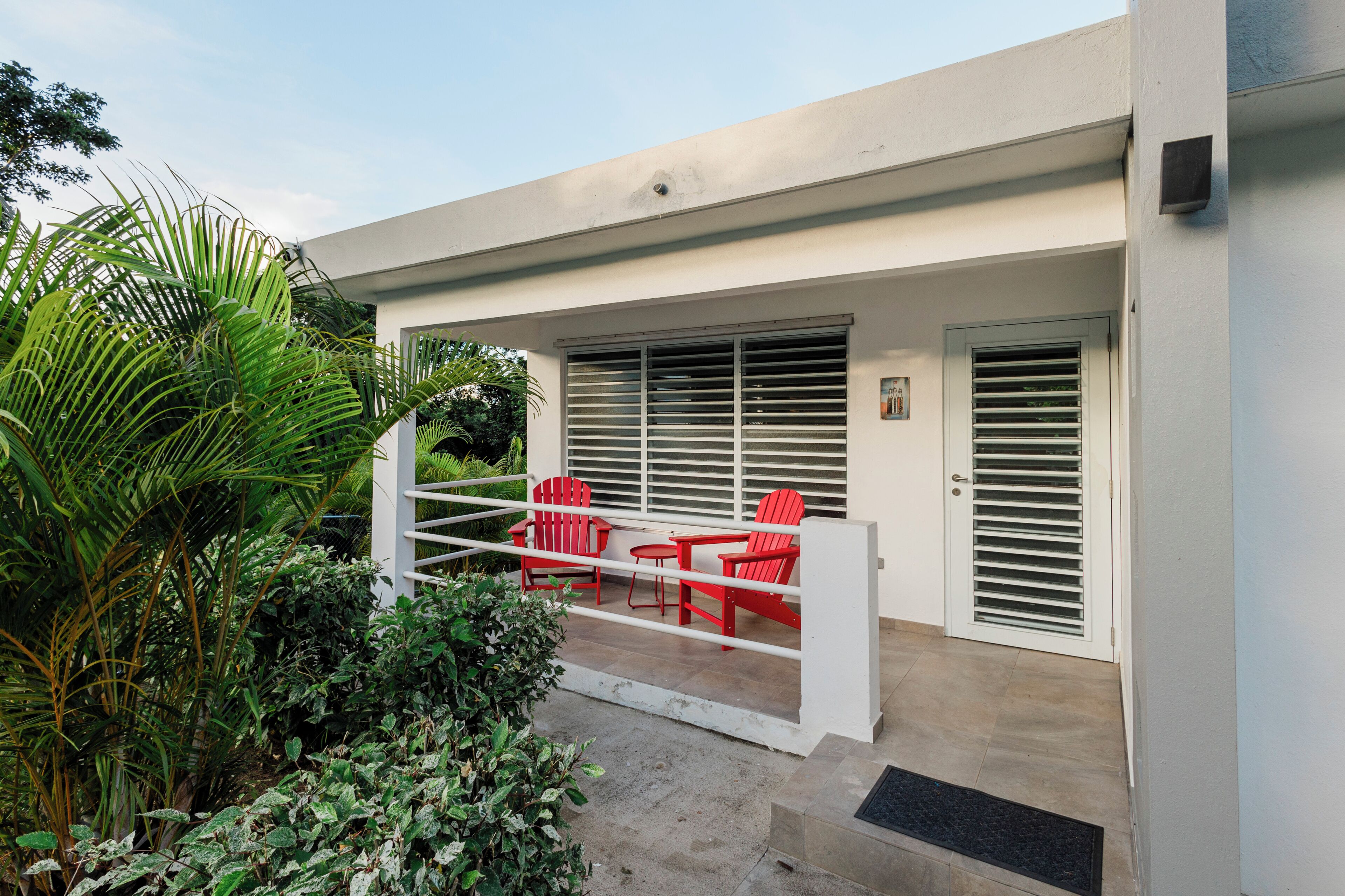 Um pátio moderno ao ar livre com guarda-corrimão branco, cadeiras vermelhas e uma porta com ripas horizontais.