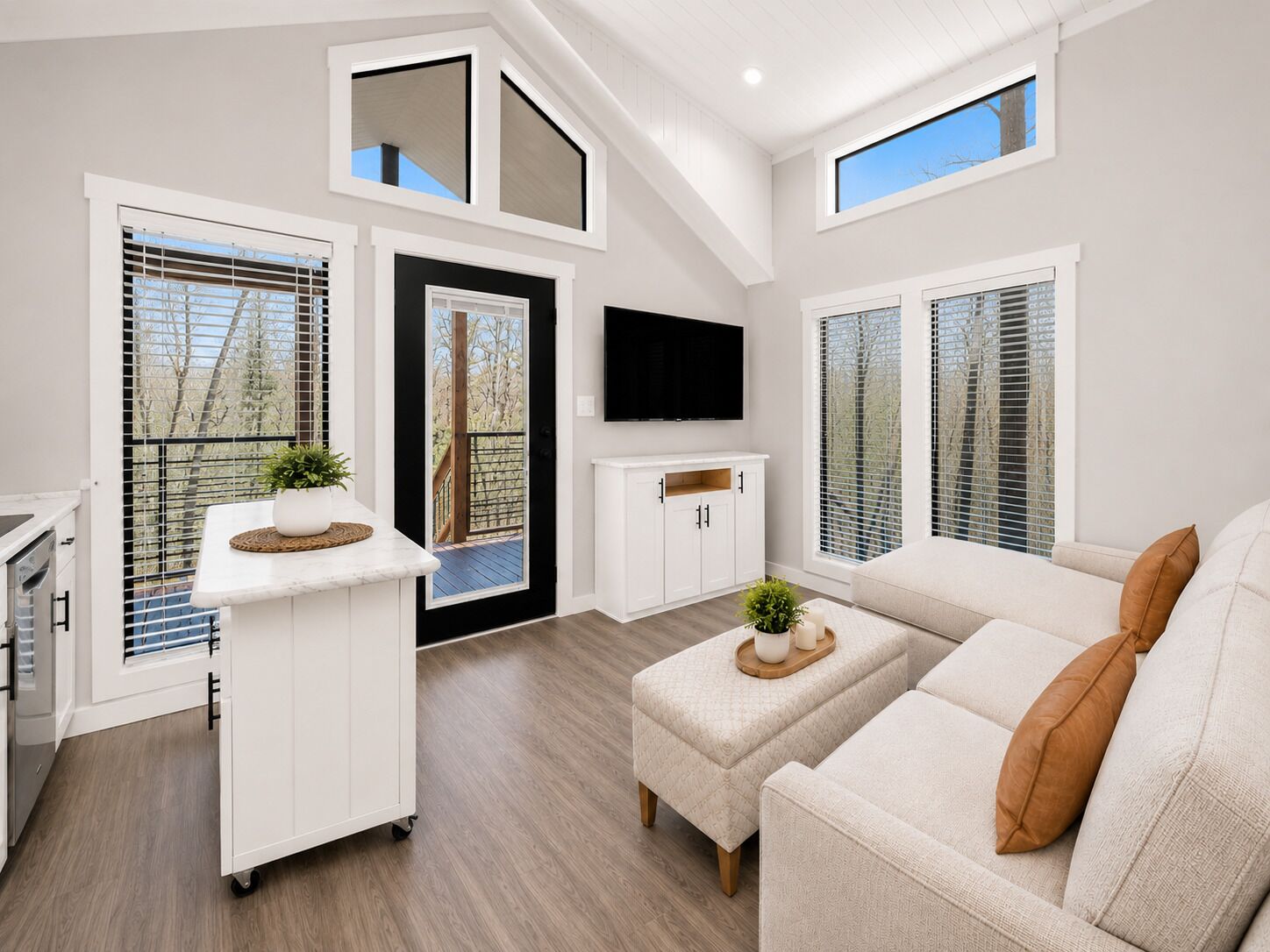 A modern living room with a sofa, a TV, a kitchen area, and a balcony with a view.