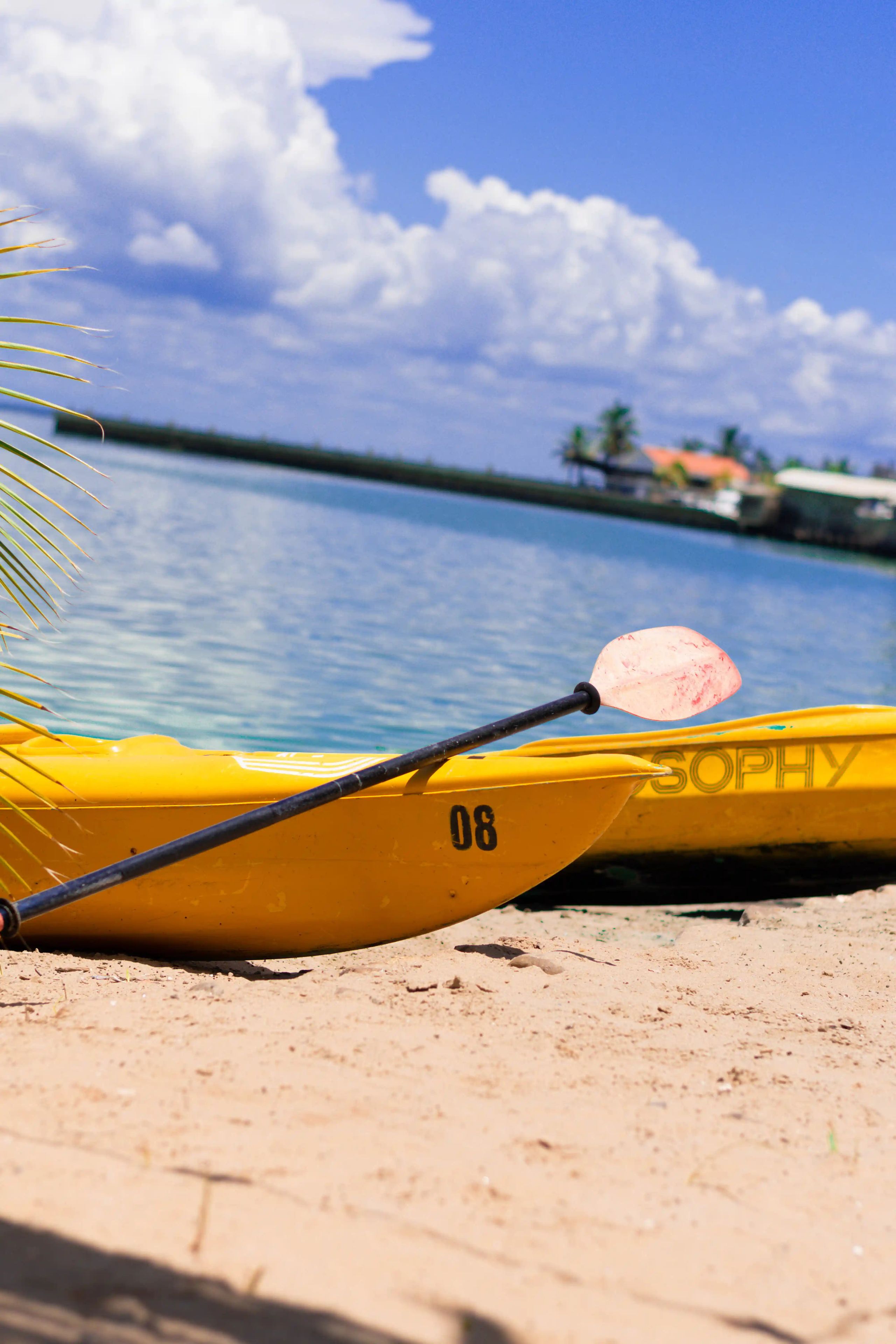 A yellow kayak with the number 06 and the word 'SOPHY' on it, resting on a sandy beach with a clear blue sky and calm water in the background.