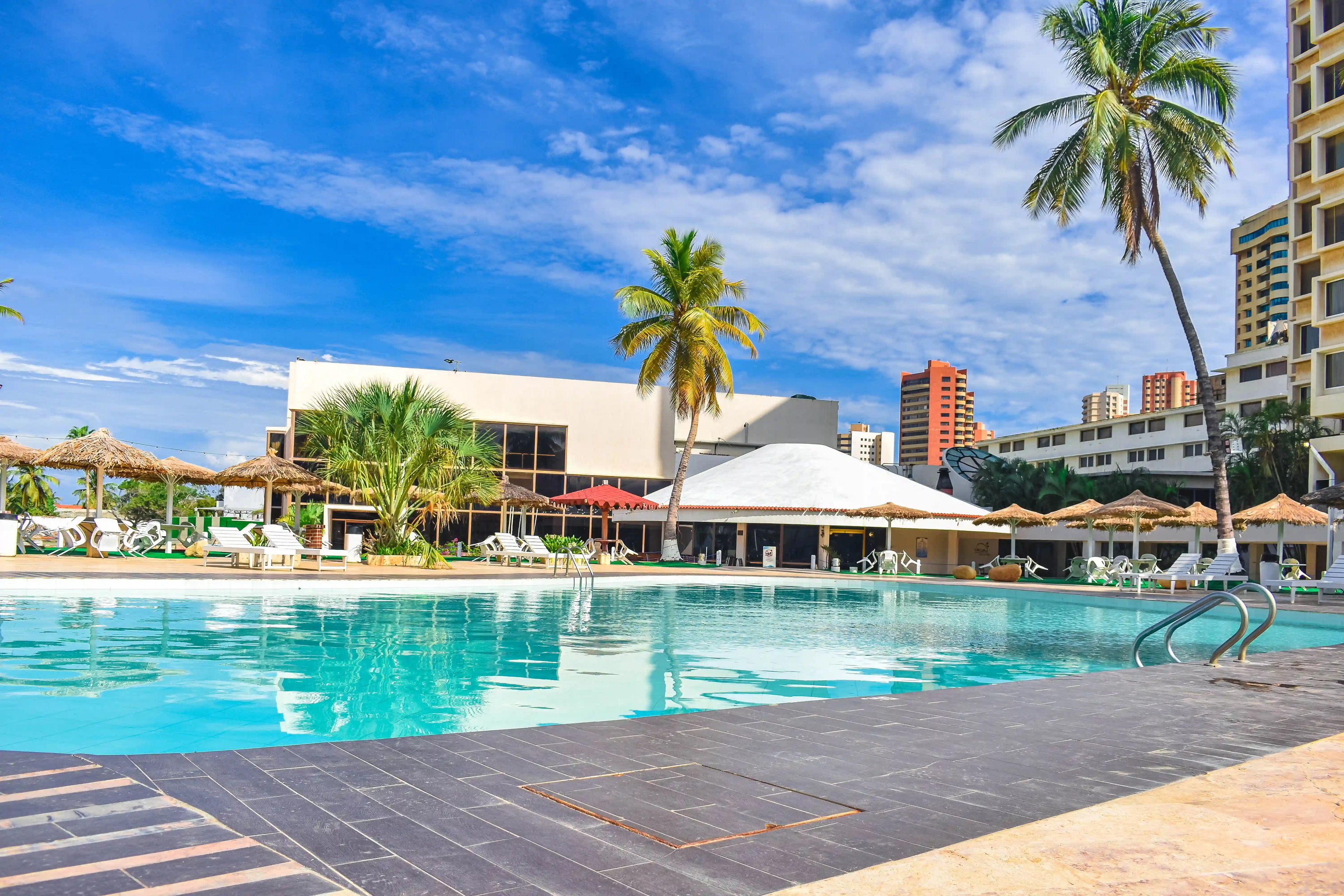 A swimming pool with lounge chairs and umbrellas, surrounded by palm trees and buildings.
