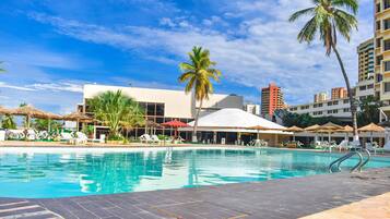 A swimming pool with lounge chairs and umbrellas, surrounded by palm trees and buildings.