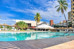A swimming pool with lounge chairs and umbrellas, surrounded by palm trees and buildings. - Hotel Tibisay Maracaibo (Maracaibo)