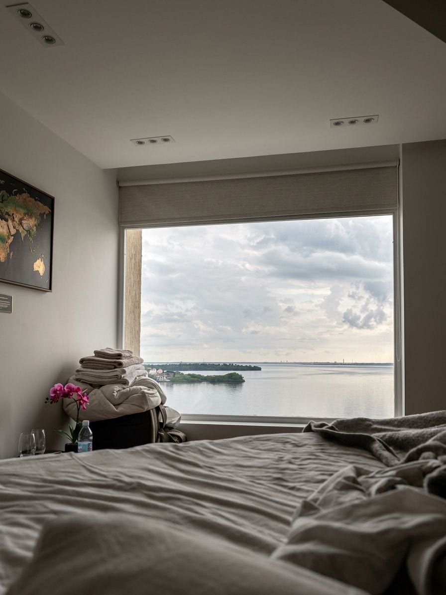 A bedroom with a large window, a bed, a bedside table with a lamp, and a map on the wall.