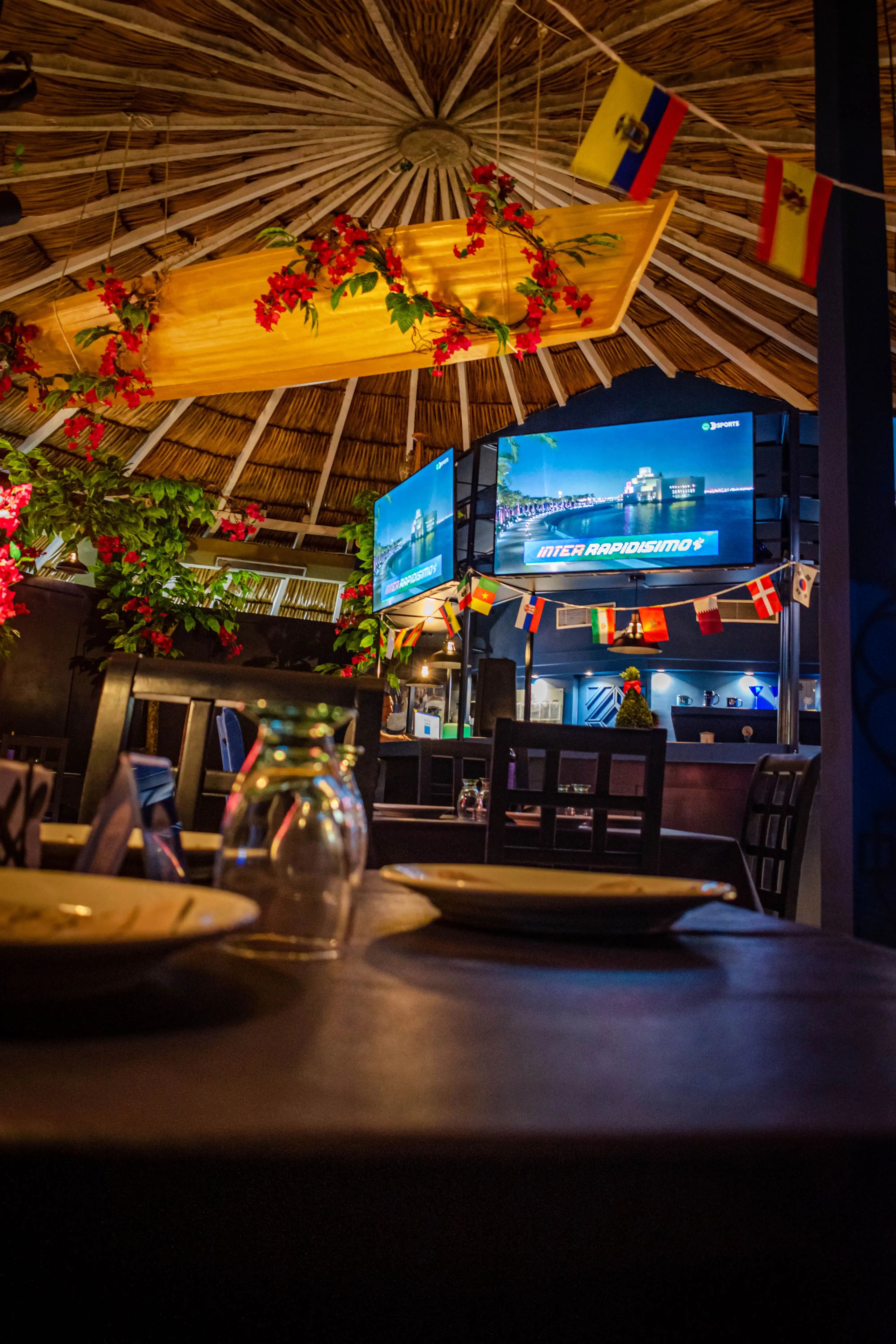 A restaurant interior with a thatched roof, multiple screens displaying sports content, and a variety of flags.